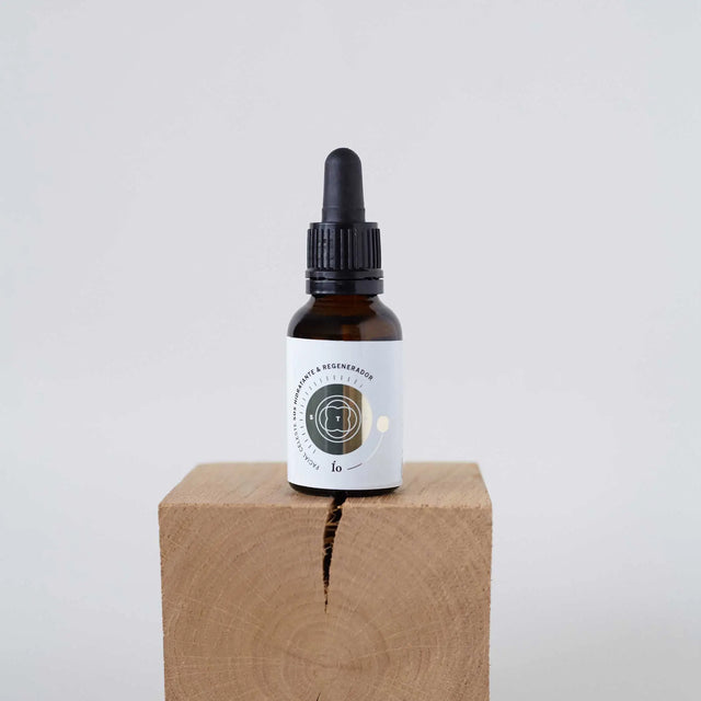 Botella de aceite o sérum con cuentagotas, con etiqueta de diseño circular que contiene texto ilegible, presentada sobre un cubo de madera con fondo blanco.