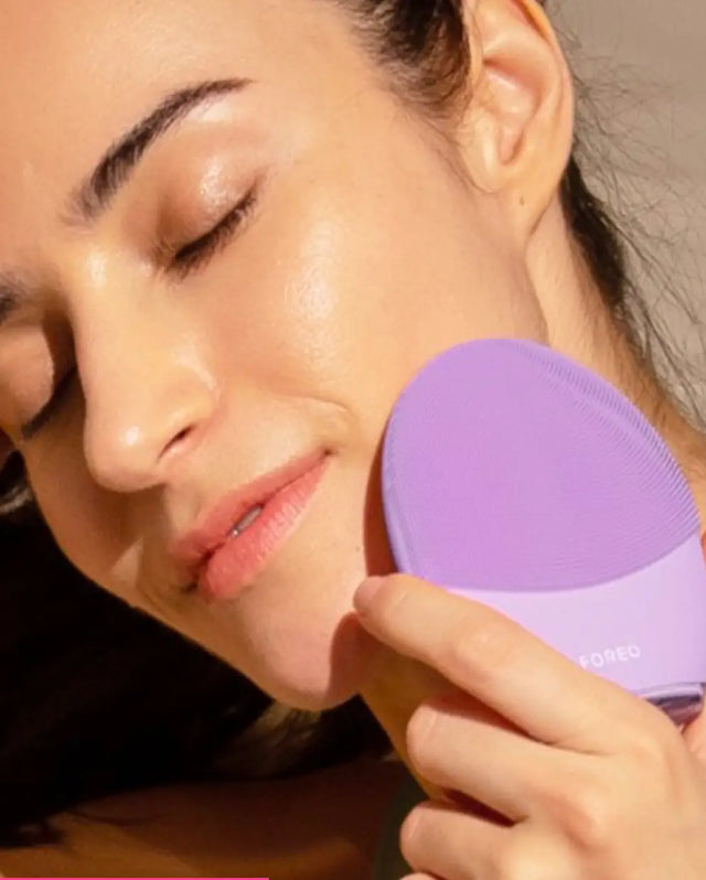 Primer plano de una mujer con los ojos cerrados, disfrutando mientras utiliza un cepillo de limpieza facial de silicona vibratorio de la marca FOREO, de color lavanda o morado claro, sobre su mejilla.