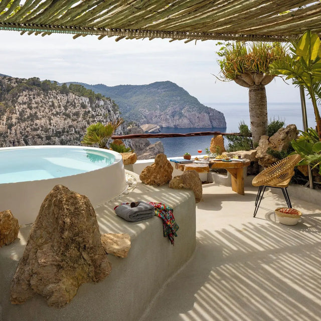 Terraza con jacuzzi circular blanco rodeado de rocas decorativas, zona de descanso con mesa rústica, sillas y bebidas, frente a acantilados verdes y el mar Mediterráneo Terraza con jacuzzi circular blanco rodeado de rocas decorativas, zona de descanso con mesa rústica, sillas y bebidas, frente a acantilados verdes y el mar Mediterráneo