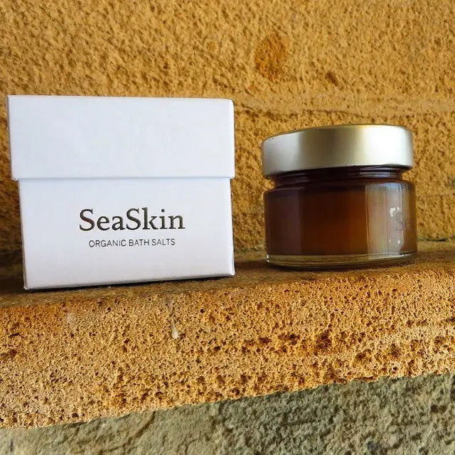 Un frasco pequeño de vidrio oscuro con tapa dorada, lleno de sales de baño o producto similar, junto a su caja de cartón blanca etiquetada como 'SeaSkin Organic Bath Salts'. Ambos productos están colocados sobre una superficie rugosa de piedra o ladrillo de color ocre o naranja. SeaSkin Honig-Gesichtsmaske