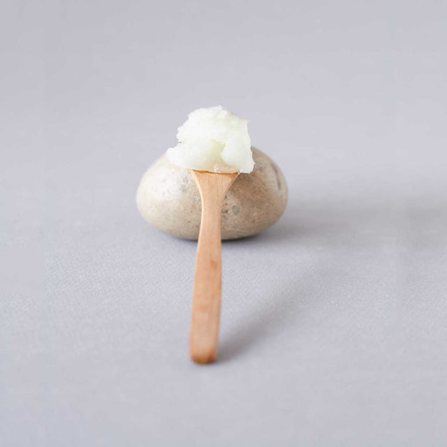 Cuchara de madera con bálsamo exfoliante corporal nutritivo de Beseaskin, 100% orgánico a base de Sándalo y Naranja sobre piedra con fondo gris.