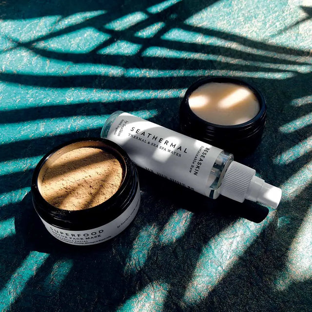 Envases de Bebalm Face Cleanser (limpiador), SuperFood Face Mask (energizante) y Tónico Seathermal (hidratante) de Beseaskin, 100% orgánicos sobre superficie azul con sombra de palmera. Anti-Akne-Set