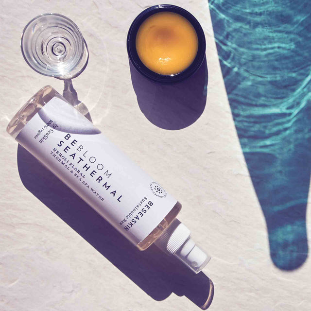 Primer plano en un ambiente soleado de un producto de belleza 'BESEA SKIN BE BLOOM SEATHERMAL' spray de Neroli Floral y Agua de Mar Termal, junto a un tarro de crema anaranjada y una copa de vidrio. El reflejo del agua de la piscina crea un patrón de luz y sombra azul