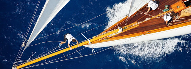 Vista aérea de un velero en movimiento con un tripulante caminando sobre el mástil, rodeado de un mar azul intenso. Vista aérea de un velero en movimiento con un tripulante caminando sobre el mástil, rodeado de un mar azul intenso.