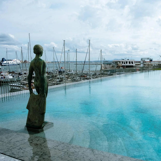 Vista desde la piscina infinita del Club de Mar en Palma de Mallorca, con una escultura femenina en primer plano y veleros atracados en el puerto al fondo bajo un cielo nublado. Vista desde la piscina infinita del Club de Mar en Palma de Mallorca, con una escultura femenina en primer plano y veleros atracados en el puerto al fondo bajo un cielo nublado.