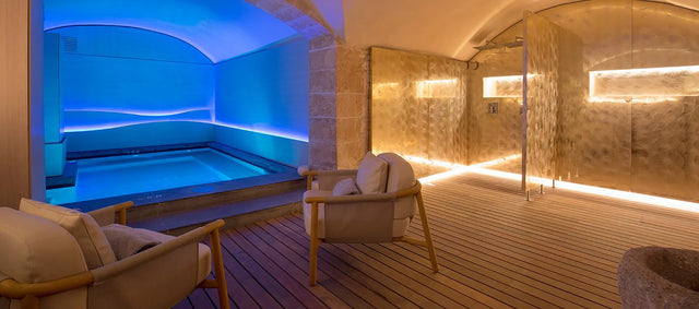 Zona de spa interior con piscina iluminada en tonos azules, paredes de piedra y duchas doradas, creando un ambiente elegante y relajante. Zona de spa interior con piscina iluminada en tonos azules, paredes de piedra y duchas doradas, creando un ambiente elegante y relajante.