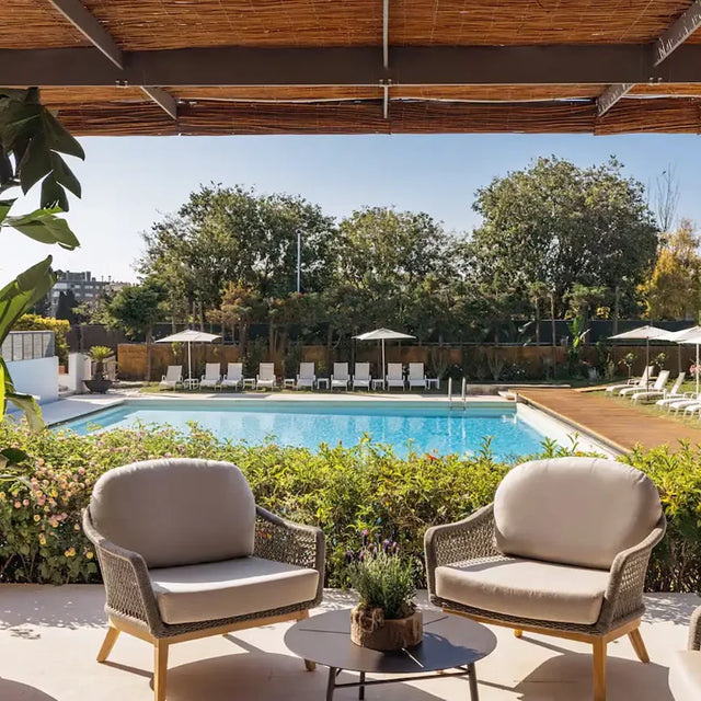 Vista de una piscina al aire libre con tumbonas alineadas y sombrillas, enmarcada por vegetación y árboles, con dos sillones acolchados y una mesa baja en primer plano bajo una pérgola Vista de una piscina al aire libre con tumbonas alineadas y sombrillas, enmarcada por vegetación y árboles, con dos sillones acolchados y una mesa baja en primer plano bajo una pérgola