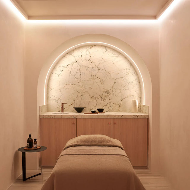 Sala de tratamiento minimalista con camilla cubierta por manta beige, pared de mármol con arco iluminado y mueble de madera clara, acompañada por una mesa auxiliar con botellas de productos Sala de tratamiento minimalista con camilla cubierta por manta beige, pared de mármol con arco iluminado y mueble de madera clara, acompañada por una mesa auxiliar con botellas de productos