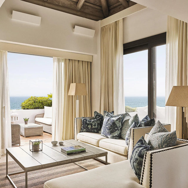 Salón luminoso con sofás claros y cojines estampados, mesa de centro de madera y lámparas de pie, con cortinas abiertas que revelan una terraza amueblada y vistas al mar Salón luminoso con sofás claros y cojines estampados, mesa de centro de madera y lámparas de pie, con cortinas abiertas que revelan una terraza amueblada y vistas al mar