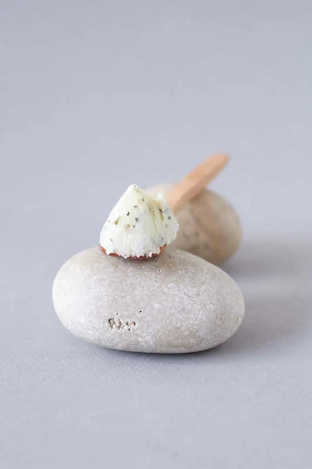 Una pequeña muestra de producto cosmético cremoso y blanco, con motas oscuras, colocada sobre una piedra de río lisa de color gris claro. Hay una segunda piedra y un aplicador de madera suavemente desenfocados detrás.