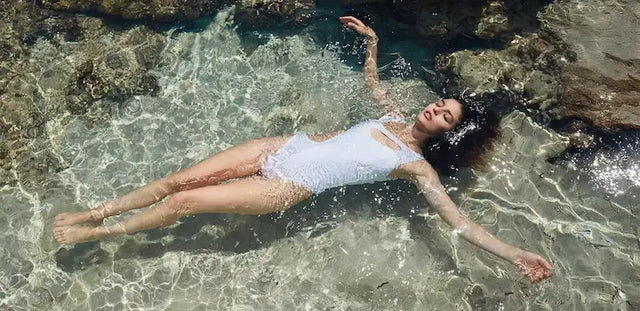 Mujer joven flotando relajadamente boca arriba en agua turquesa cristalina en playa rocosa, vistiendo traje de baño entero blanco de tirantes.