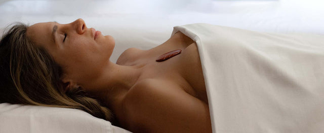 Mujer con los ojos cerrados, cubierta por una toalla blanca, recibiendo un tratamiento de spa con una piedra o cristal curativo sobre el pecho. Relajación y bienestar.