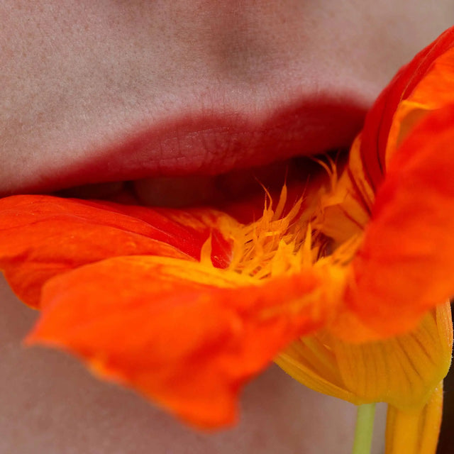 Primer plano de labios con labial mate color rojo anaranjado tocando pétalos de una flor naranja brillante. Representa cosméticos naturales, maquillaje labios vibrante o ingredientes florales para la piel. BLOOSOM TO GLOW RITUAL | 120 MIN