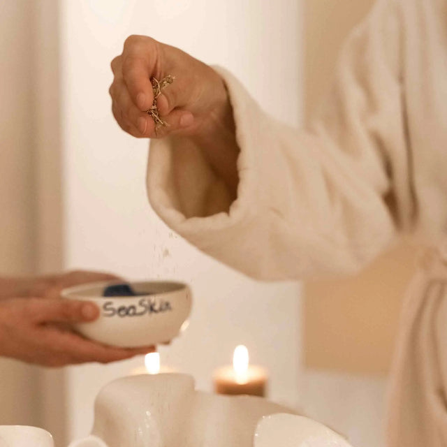 Persona con albornoz blanco de spa espolvoreando hierbas secas en un recipiente sobre velas encendidas. Proceso de tratamiento corporal o facial de lujo con ingredientes naturales. ALQUIMISTISCHE KÖRPERMASSAGE | Can Marqués Palma