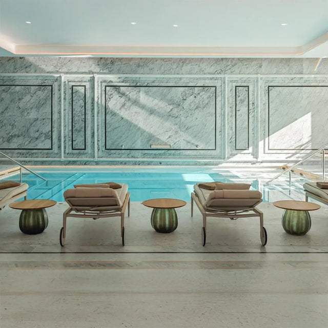 Piscina interior con paredes de mármol gris, camillas acolchadas alineadas y mesas auxiliares redondas, iluminada por luz natural que entra desde el techo Piscina interior con paredes de mármol gris, camillas acolchadas alineadas y mesas auxiliares redondas, iluminada por luz natural que entra desde el techo