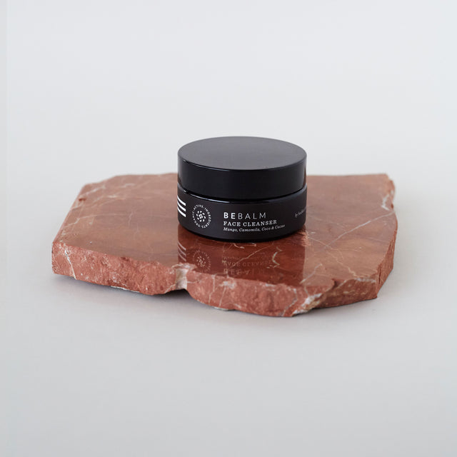 Envase de bálsamo facial desmaquillante de Beseaskin, 100% orgánico a base de manteca de mango y cacao, sobre piedra en una mesa blanca.