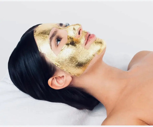 Mujer recibiendo un tratamiento facial de lujo, con su rostro cubierto por una máscara de pan de oro, reclinada sobre una toalla blanca.