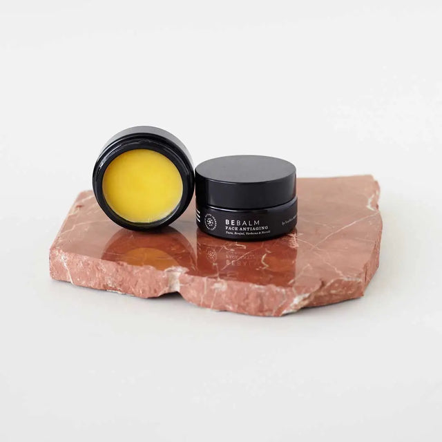 Dos pequeños tarros de bálsamo facial antiedad 'BE BALM' de color negro. Uno está abierto, mostrando el contenido amarillo brillante del bálsamo. Los tarros están colocados sobre una losa de piedra de color terracota o marrón rojizo irregular.