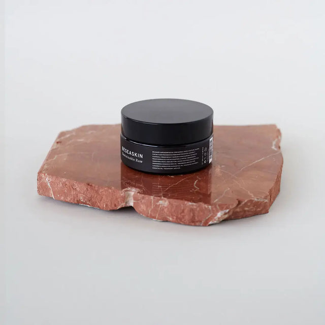 Envase de bálsamo facial desmaquillante de Beseaskin, 100% orgánico a base de manteca de mango y cacao, sobre piedra en una mesa blanca.