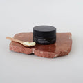Tarro cerrado de bálsamo limpiador facial 'BE BALM' de color negro, sobre una losa de piedra de color terracota o marrón rojizo irregular. Una pequeña cucharada de bálsamo cremoso de color amarillo pálido se apoya en una espátula de madera a la izquierda.