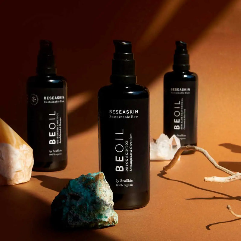 Envases de aceite corporal reafirmante de Beseaskin, 100% orgánico a base de Lemongrass y Geranio con fondo naranja y piedras alrededor.