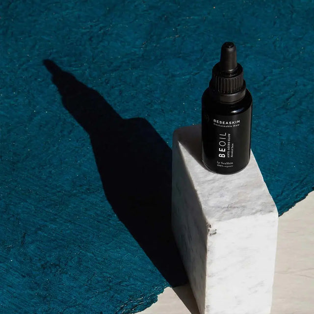 Frasco gotero (dropper) oscuro de sérum o aceite facial 'BE OIL' de la marca SEASKIN, colocado sobre un bloque de mármol o piedra blanca, proyectando una larga sombra sobre un fondo texturizado de color azul turquesa profundo.