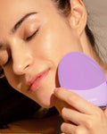 Primer plano de una mujer con los ojos cerrados, disfrutando mientras utiliza un cepillo de limpieza facial de silicona vibratorio de la marca FOREO, de color lavanda o morado claro, sobre su mejilla.