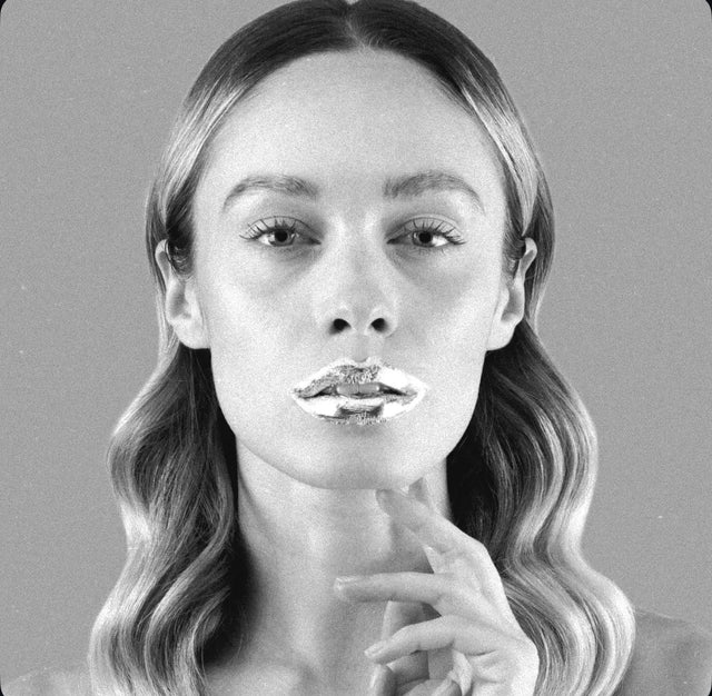 Retrato de belleza en blanco y negro de una mujer con maquillaje dramático en los labios con efecto plateado o metalizado. Tiene una mano tocando suavemente la barbilla.