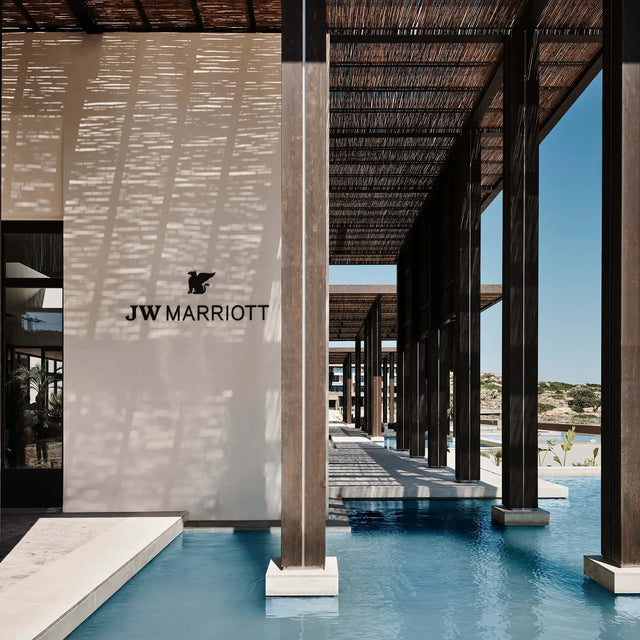 Entrada exterior del hotel JW Marriott con columnas de madera sobre piscina poco profunda, pérgola de cañas proyectando sombras sobre la fachada blanca y cielo azul despejado de fondo