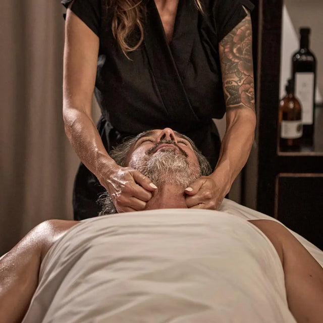 Hombre con barba recibiendo un masaje y tratamiento facial para hombres en el cuello y mandíbula. Esteticista aplicando técnica profesional en el spa PURO PALACIO CAN MARQUÉS.