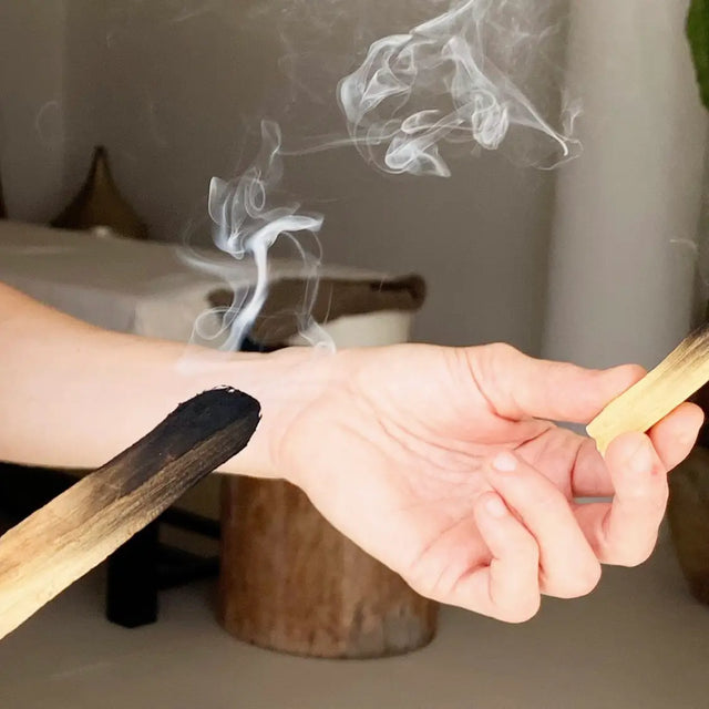 Manos sujetando dos piezas de Palo Santo prendidas, con humo blanco.