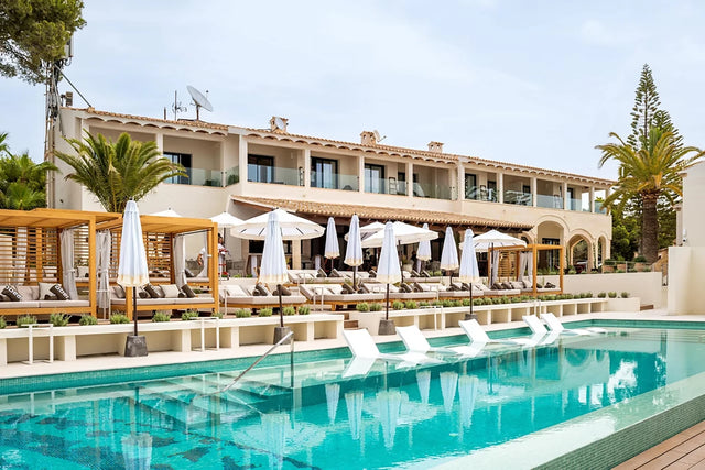 Vista exterior del PuroBeach Resort Santa Ponsa, con su arquitectura mediterránea, balcones con barandillas de vidrio y una piscina rodeada de camas balinesas y sombrillas blancas.