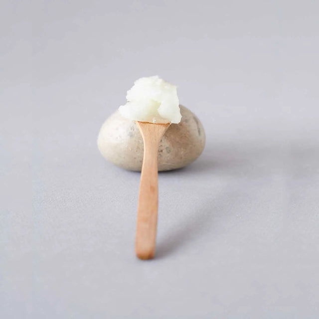 Cuchara de madera con bálsamo exfoliante corporal nutritivo de Beseaskin, 100% orgánico a base de Sándalo y Naranja sobre piedra con fondo gris.