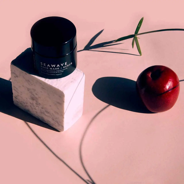Tarro de bálsamo o mascarilla facial de color negro, con etiqueta blanca de la marca SEAWAVE, colocado sobre un pequeño bloque de mármol blanco. Hay una manzana roja y un tallo de planta verde al lado, con sombras largas sobre un fondo rosa pálido.