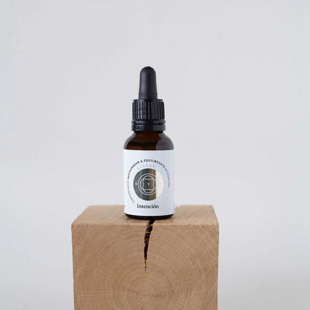 Frasco gotero (dropper) de vidrio ámbar, probablemente un sérum o aceite, con una pequeña etiqueta circular, colocado sobre un bloque de madera clara con una grieta visible, con un fondo blanco liso.