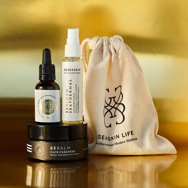Envases de Bebalm Face Cleanser (limpiador), Bebloom Seathermal Lavanda (calmante) y Elixir Ío (hidratante y regenerador), con bolsa de tela de Seaskin Life sobre superficie dorada.