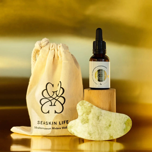 Envase de Elixir Ío (aceite hidratante y regenerador) y piedra Guasha con bolsa de tela de Seaskin Life sobre superficie dorada.