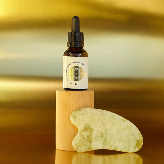 Envase de Elixir Ío (aceite hidratante y regenerador) y piedra Guasha sobre superficie y fondo dorado.
