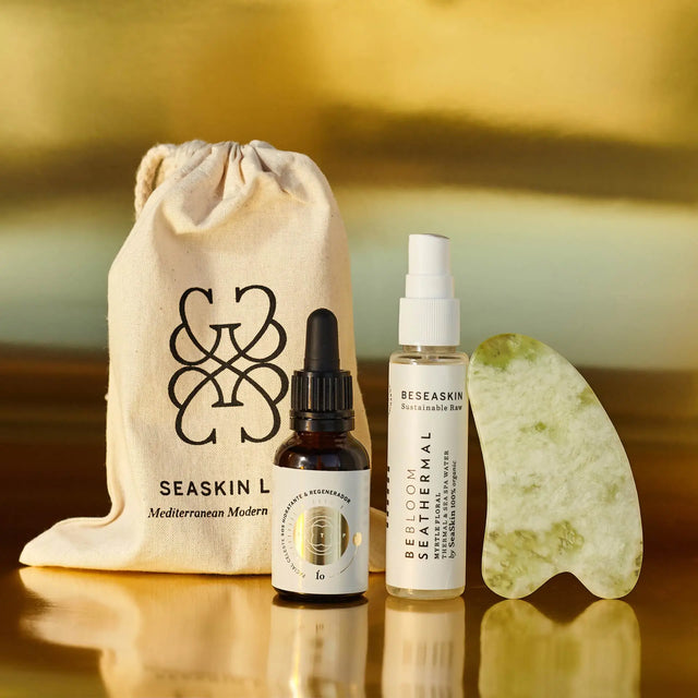 Envases de Elixir Ío (hidratante y regenerador) y Bebloom Seathermal Mirto (hidratante) junto a piedra Guasha y bolsa de tela de Seaskin Life sobre superficie dorada.