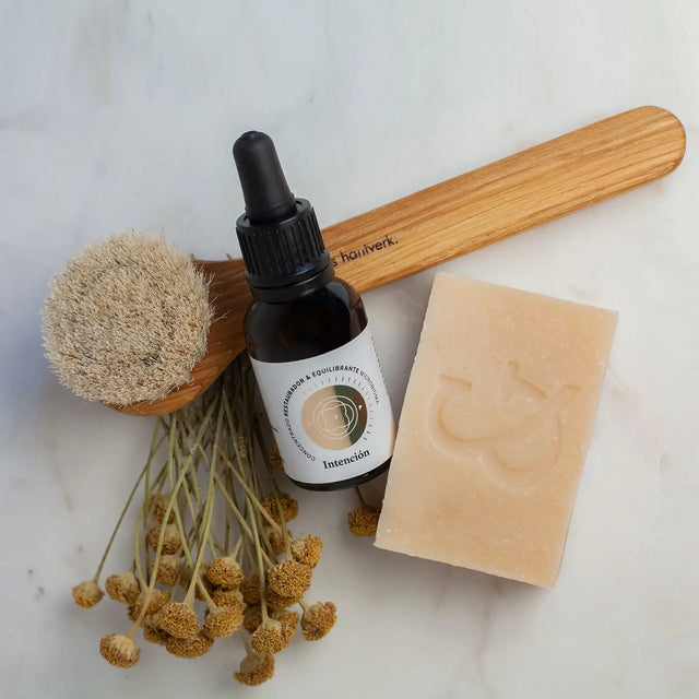 Envase de Elixir Intención (Skinbooster) junto a Jabon Orgánico Artesanal (regenerador) y Cepillo Facial de roble y pelo de cabra, 100% orgánicos sobre tabla de mármol.
