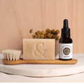 Envase de Elixir Intención (Skinbooster) junto a Jabon Orgánico Artesanal (regenerador) y Cepillo Facial de roble y pelo de cabra, 100% orgánicos sobre tabla de mármol.