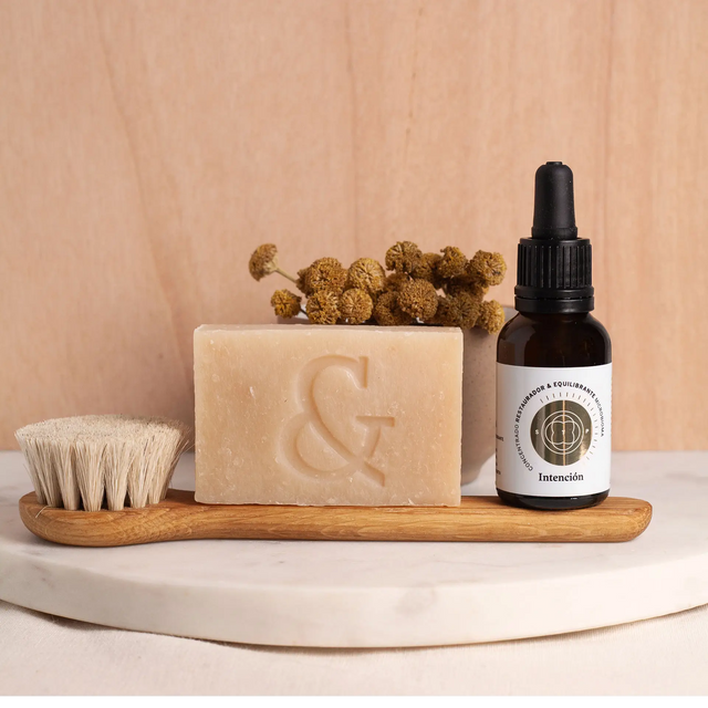 Envase de Elixir Intención (Skinbooster) junto a Jabon Orgánico Artesanal (regenerador) y Cepillo Facial de roble y pelo de cabra, 100% orgánicos sobre tabla de mármol.