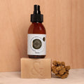 Envase de Elixir Tierra (aceite antioxidante) encima de Jabón Orgánico Artesanal (regenerador), 100% orgánicos sobre mesa de mármol con flores.