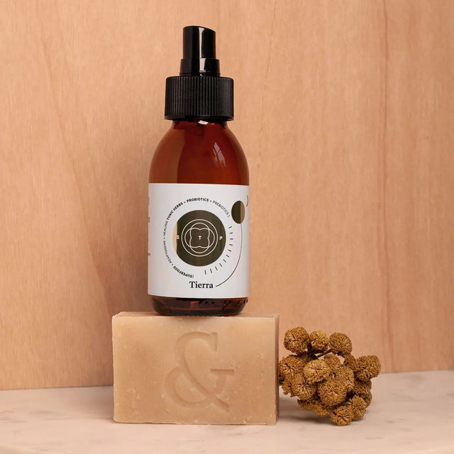 Envase de Elixir Tierra (aceite antioxidante) encima de Jabón Orgánico Artesanal (regenerador), 100% orgánicos sobre mesa de mármol con flores.