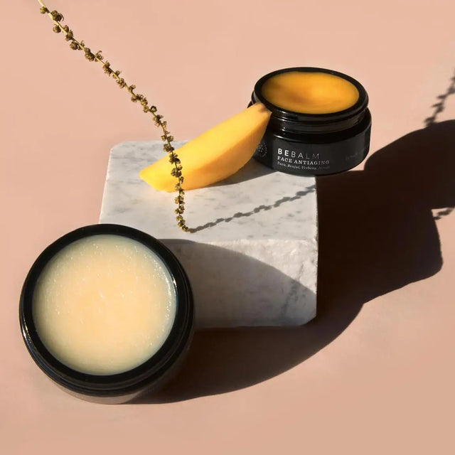 Envases de Bebalm Face Cleanser y Bebalm Face Antiaging Glow de Beseaskin, 100% orgánicos, sobre una piedra con fruta en un fondo rosa pastel.