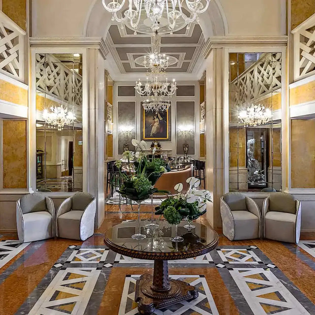 Lobby elegante con suelos de mármol, lámparas de araña colgantes, espejos altos y una mesa central con arreglos florales, en un ambiente clásico y lujoso.