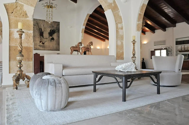 Sala de estar elegante con sofá y sillones en tonos neutros, mesa de centro oscura con escultura de coral blanco, decoración clásica con arcos de piedra, techo de vigas de madera y esculturas de caballos