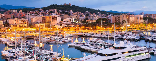 Vista del puerto Club de Mar en Palma de Mallorca al atardecer, con numerosos yates y veleros atracados, edificios iluminados al fondo y el castillo de Bellver en la cima de una colina.