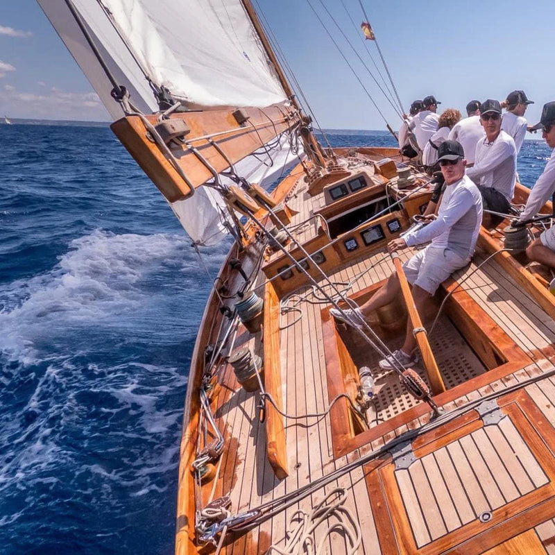 Tripulación navegando en un velero clásico de madera durante una regata en aguas abiertas, con velas desplegadas y cielo despejado.