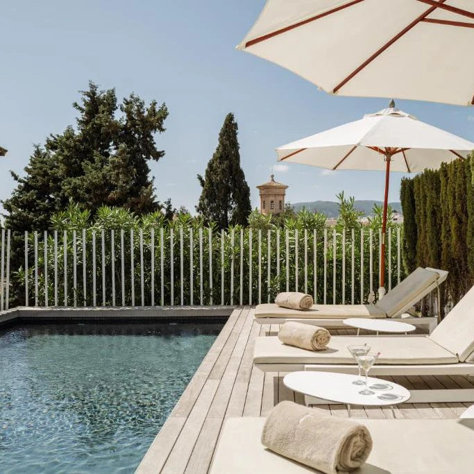 Terraza con piscina al aire libre, tumbonas con toallas enrolladas y sombrillas blancas, rodeada de vegetación y con vistas a una torre histórica en el fondo.